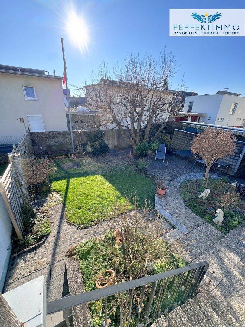 Blick von oben auf den sonnigen Gartenbereich mit Rasenfläche und gepflasterten Wegen.