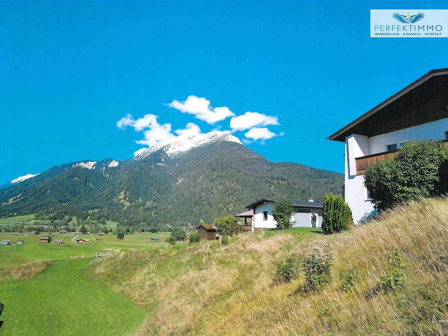Haus am Hang mit Balkon und beeindruckendem Blick auf schneebedeckte Berge und grüne Wiesen.