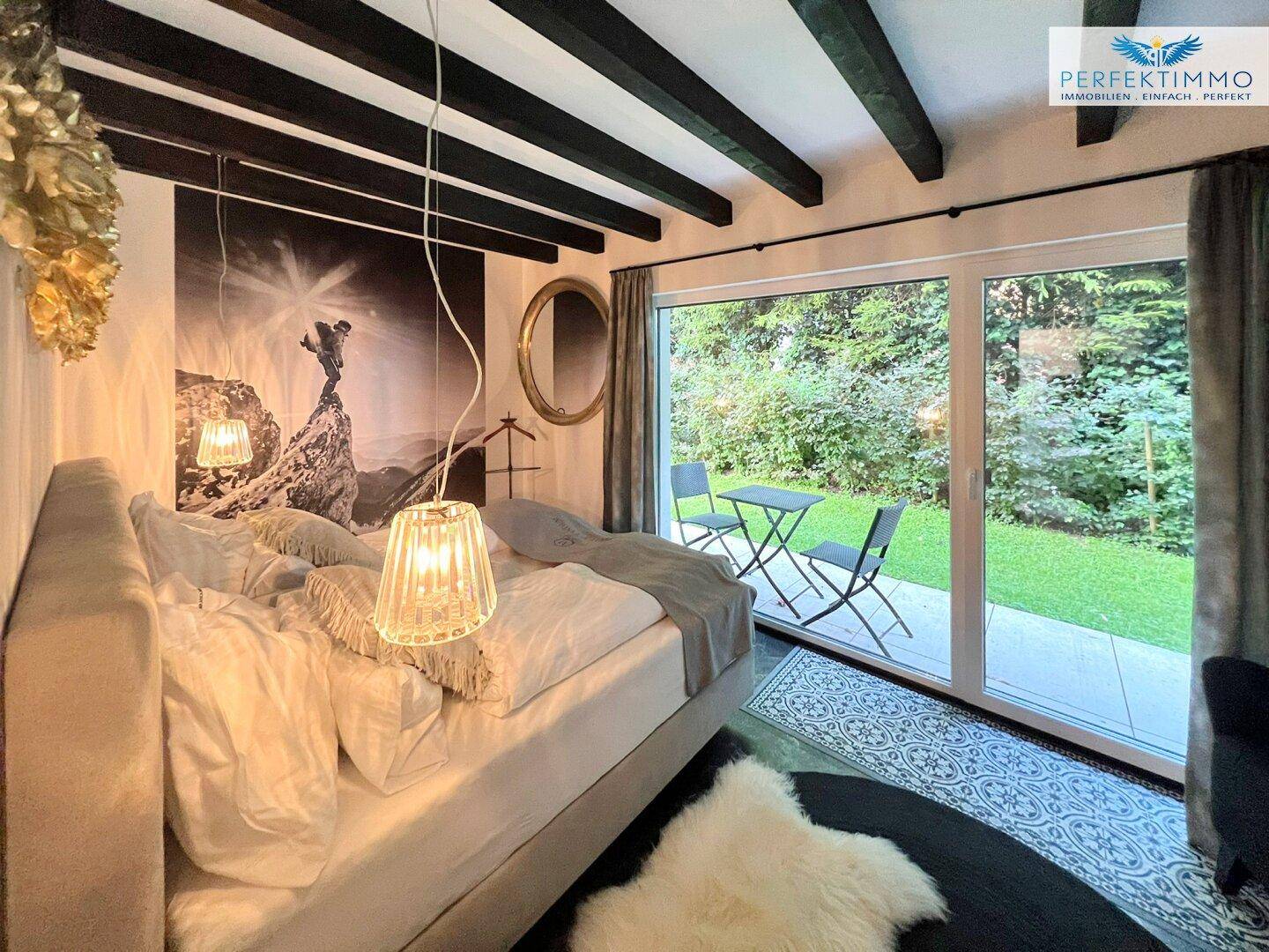 Schlafzimmer mit großem Bett, Holzbalken und direktem Zugang zu einer kleinen Terrasse im Grünen.