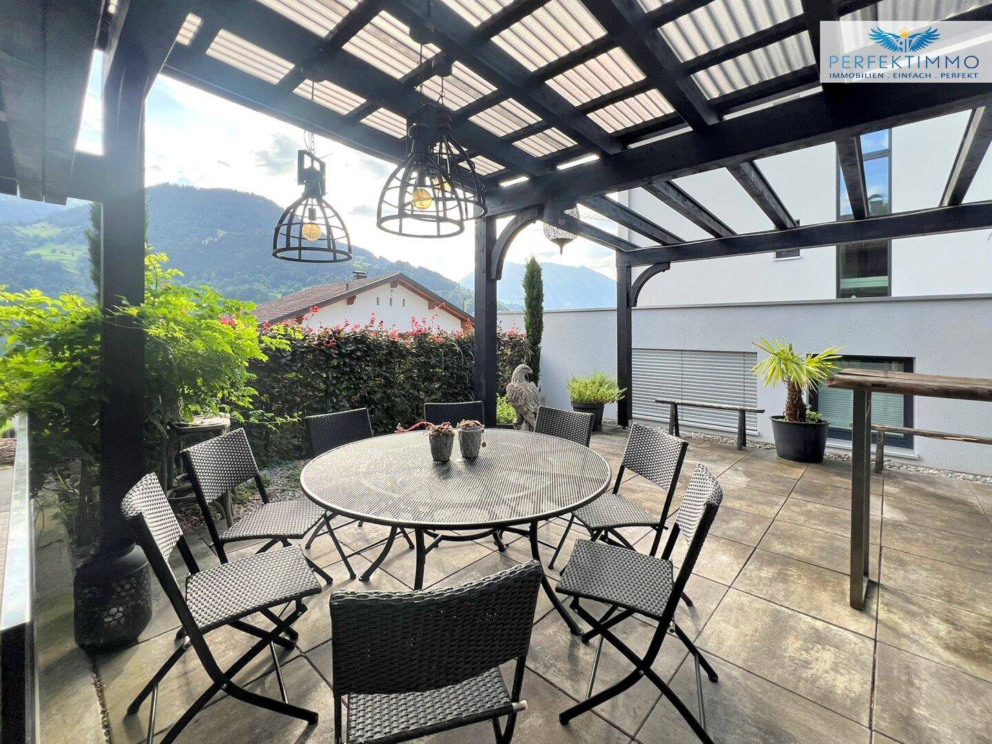 Überdachte Terrasse mit Esstisch und Stühlen, umgeben von Grün.