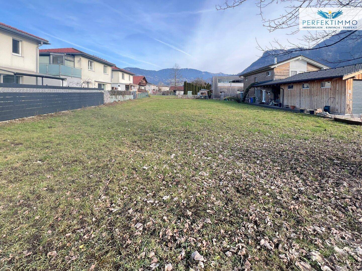 Grünes Baugrundstück mit Blick auf umliegende Wohnhäuser und die entfernten Berge.