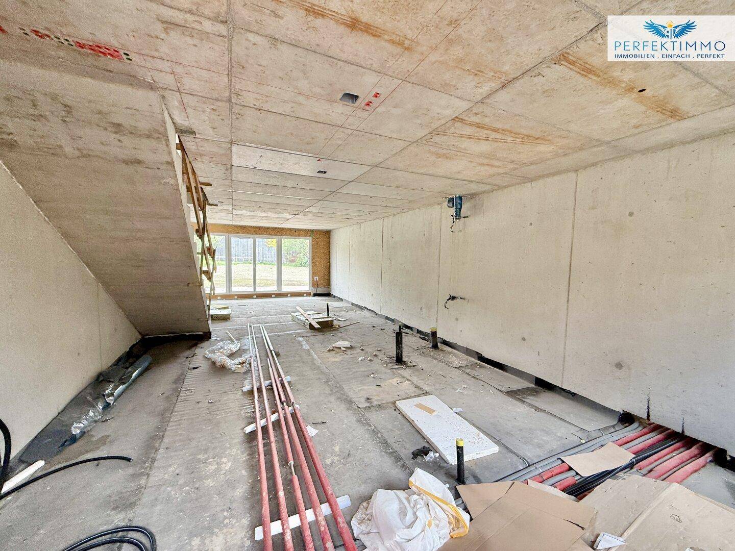 Unfertiger Innenraum im Rohbau mit Betonwänden, hohen Decken und großen Fenstern zum Garten.