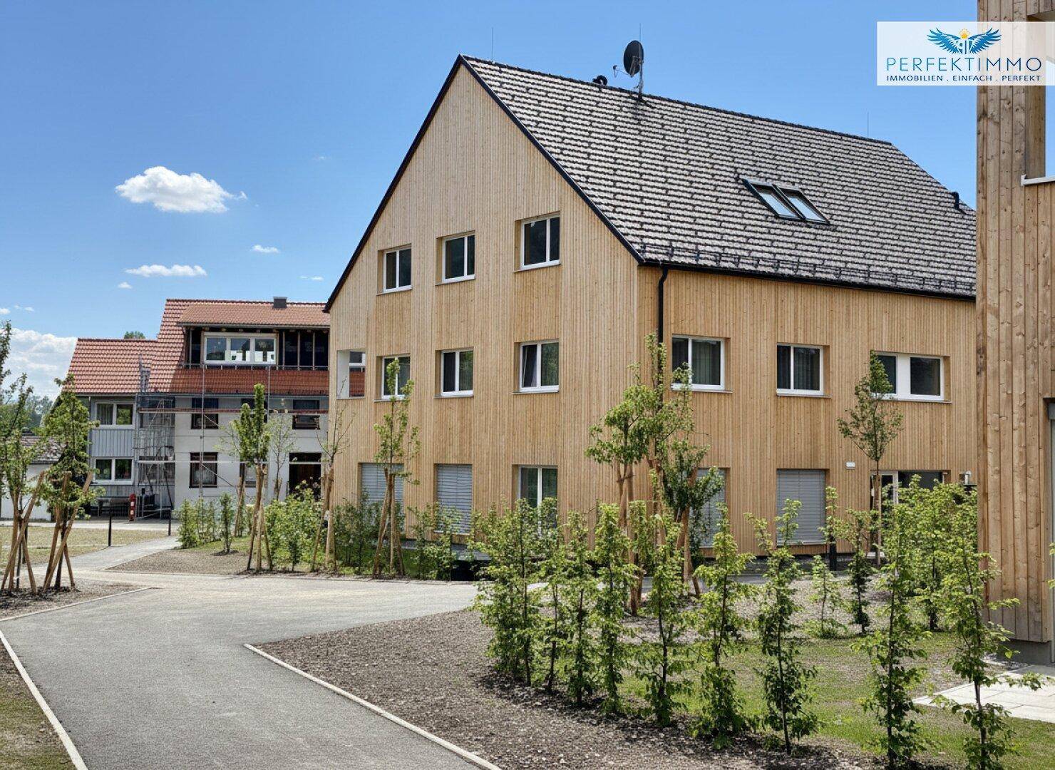 Modernes Mehrfamilienhaus mit Holzverkleidung, vielen Fenstern und grüner Umgebung.