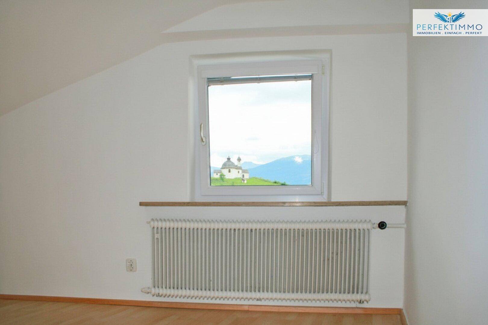 Dachzimmer mit Holzboden, weiß gestrichenen Wänden und Fenster mit Fernblick auf eine Kirche.
