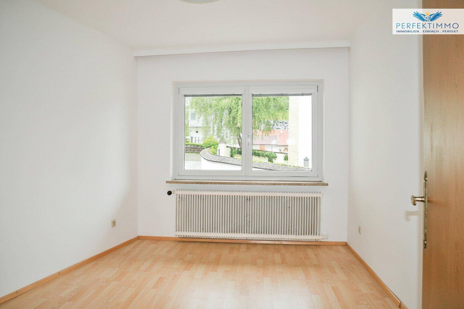 Helles Zimmer mit Holzboden, weißen Wänden und Fenster mit Blick auf Bäume.