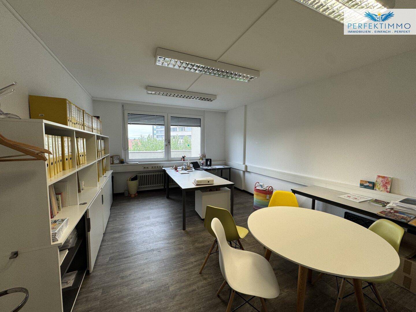 Büro mit Schreibtischen, Regalen und einem Besprechungsbereich, hell durch große Fenster.