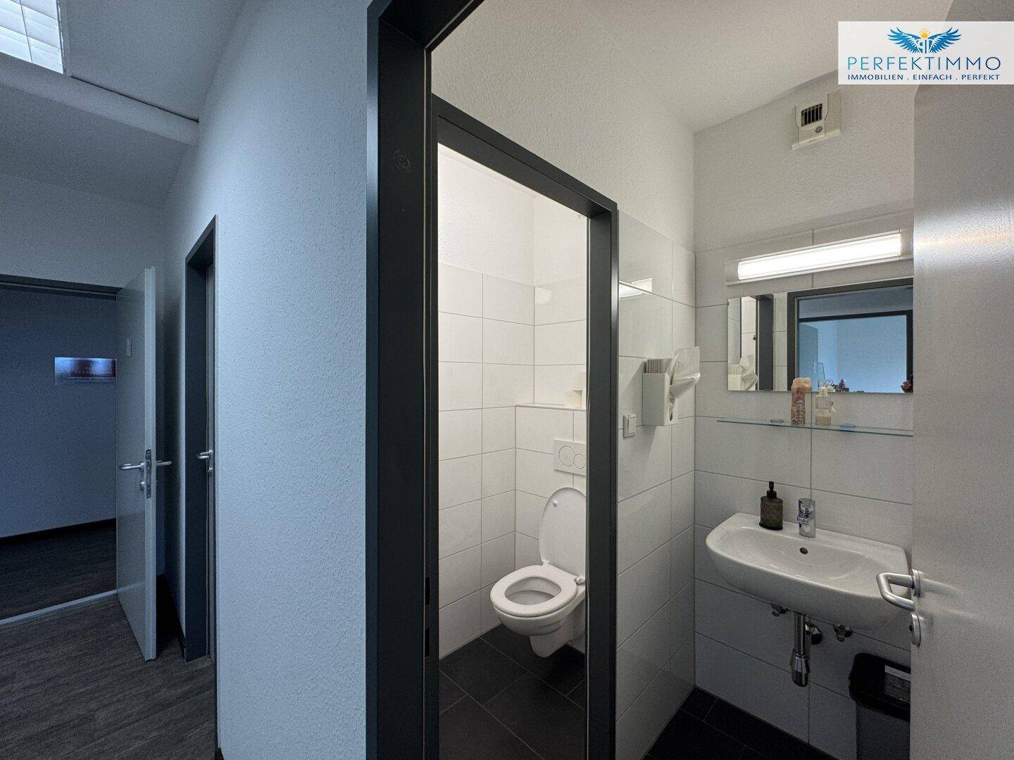 Modernes WC mit weißen Fliesen, Waschbecken und Wand-WC, gut beleuchtet und gepflegt.