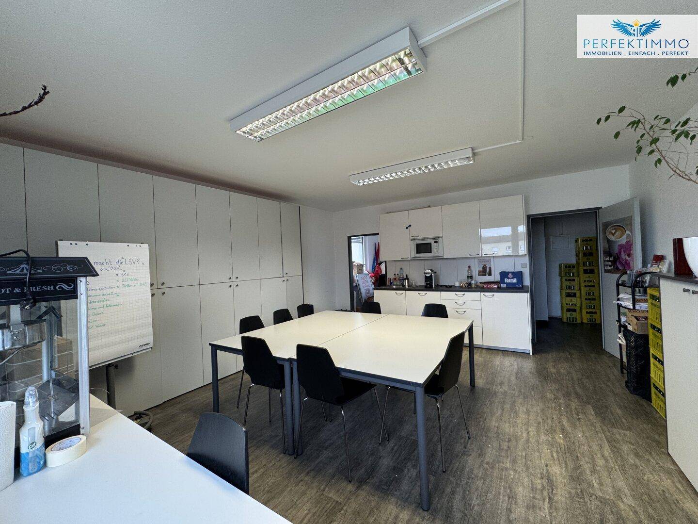 Moderne Büroküche mit weißen Hochglanzschränken, Mikrowelle und einem großen Besprechungstisch.