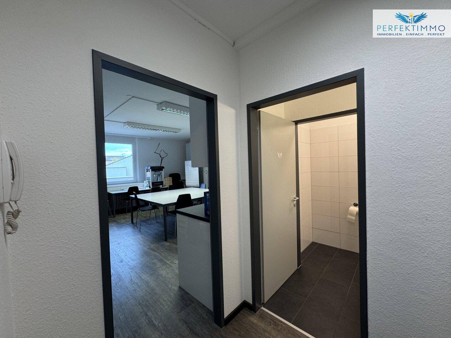 Flur mit Zugang zu einem Büro und einer Toilette, gekennzeichnet durch helle Wände und Laminatboden.