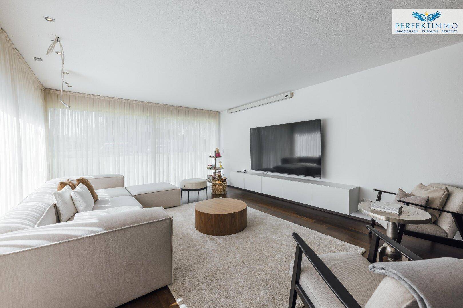 Geräumiges Wohnzimmer mit großem Sofa, Holztisch und Wandfernseher, hell durch große Fenster.