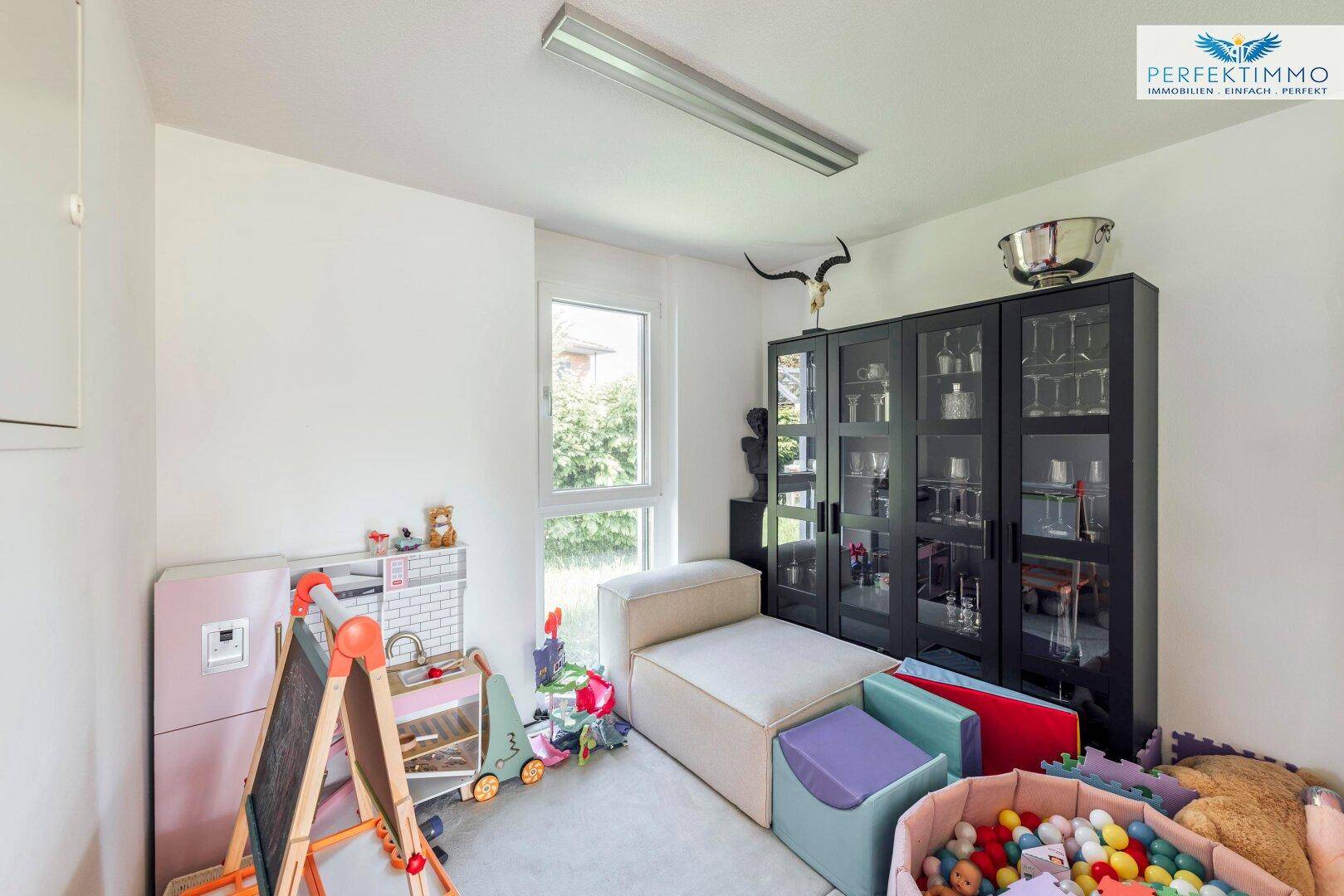 Helles Kinderzimmer mit Spielzeug, kleiner Liege und einem schwarzen Vitrinenschrank.