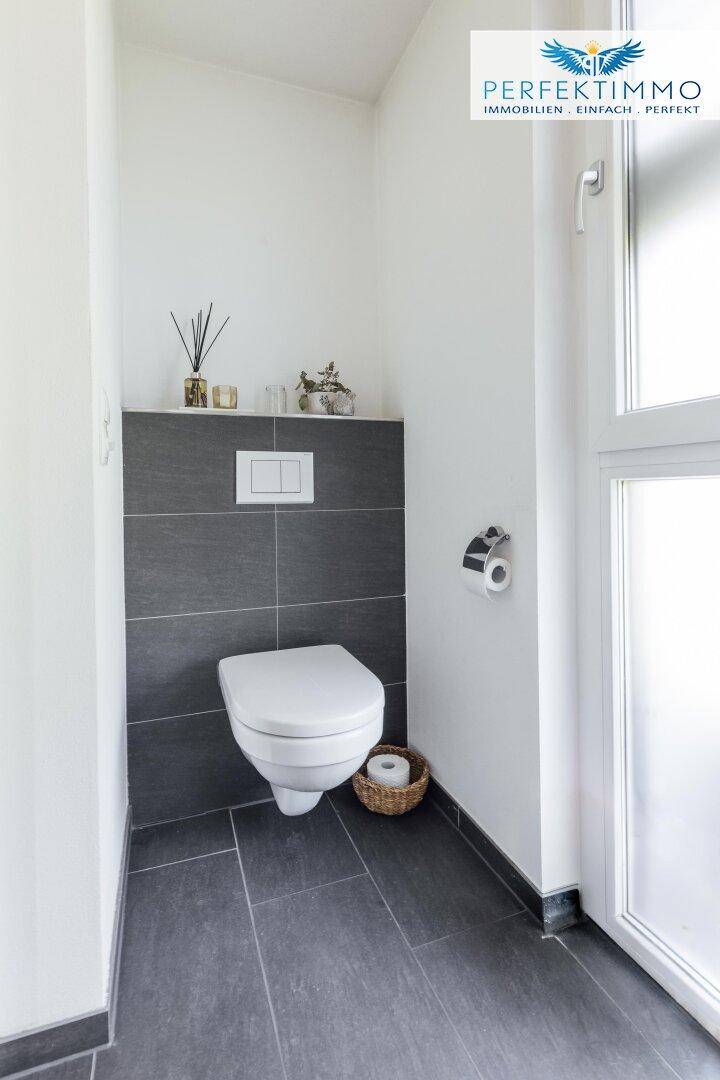 Modernes Gäste-WC mit wandhängender Toilette, dunklen Fliesen und einem Fenster für Tageslicht.