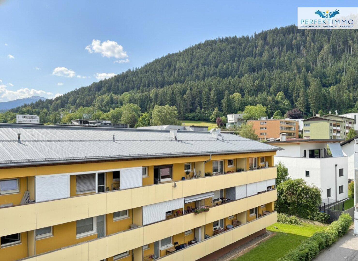 Gelbes Mehrfamilienhaus mit Balkonen, umgeben von üppigem Grün und einer bergigen Landschaft.