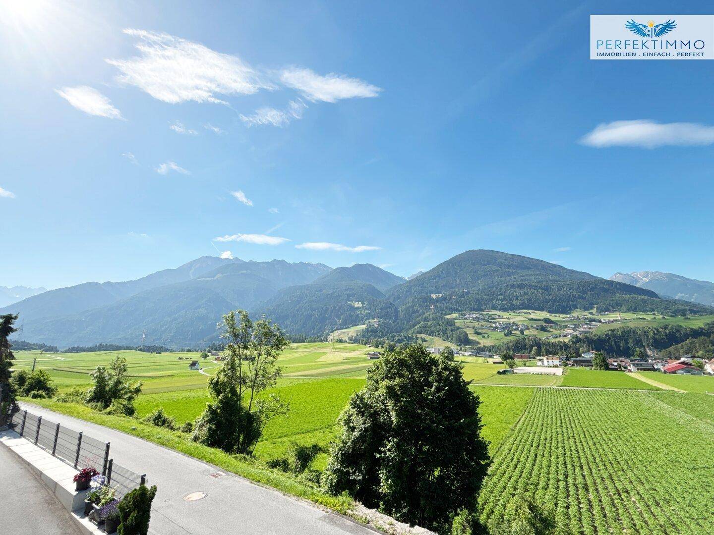 Weitläufiger Blick über grüne Felder und die majestätische Berglandschaft unter blauem Himmel.