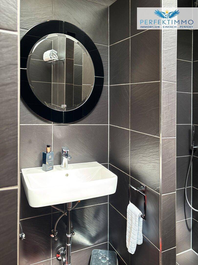 Modernes Badezimmer mit dunklen Fliesen, einem Waschbecken und einem runden Spiegel.