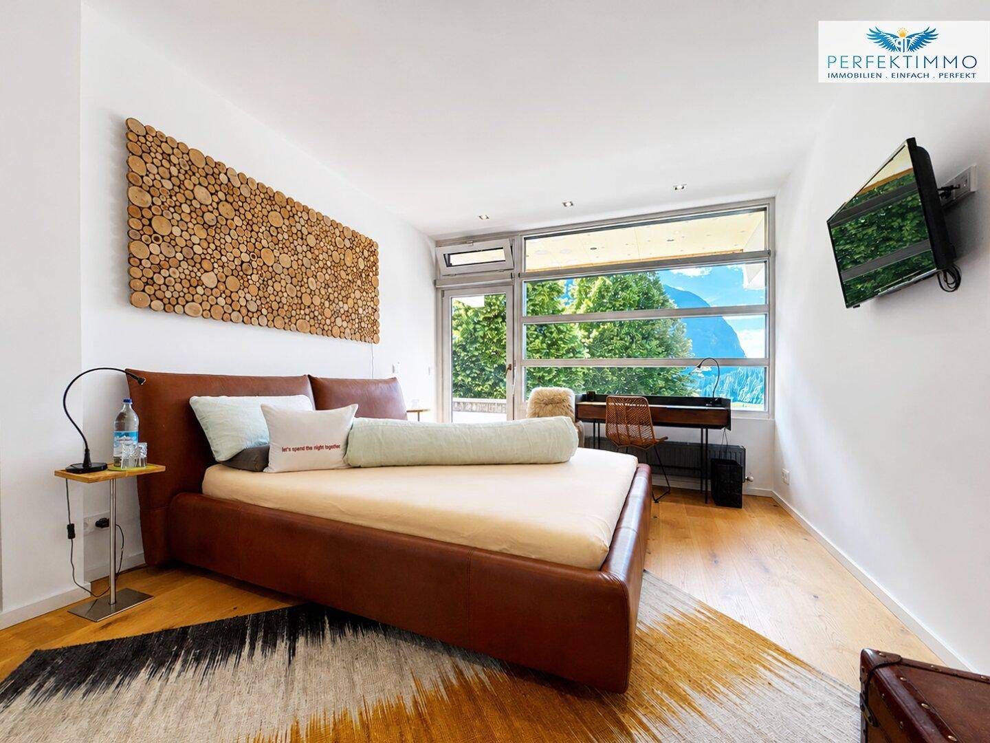 Gemütliches Schlafzimmer mit braunem Bett, Holzboden und Bergblick durch große Fenster.