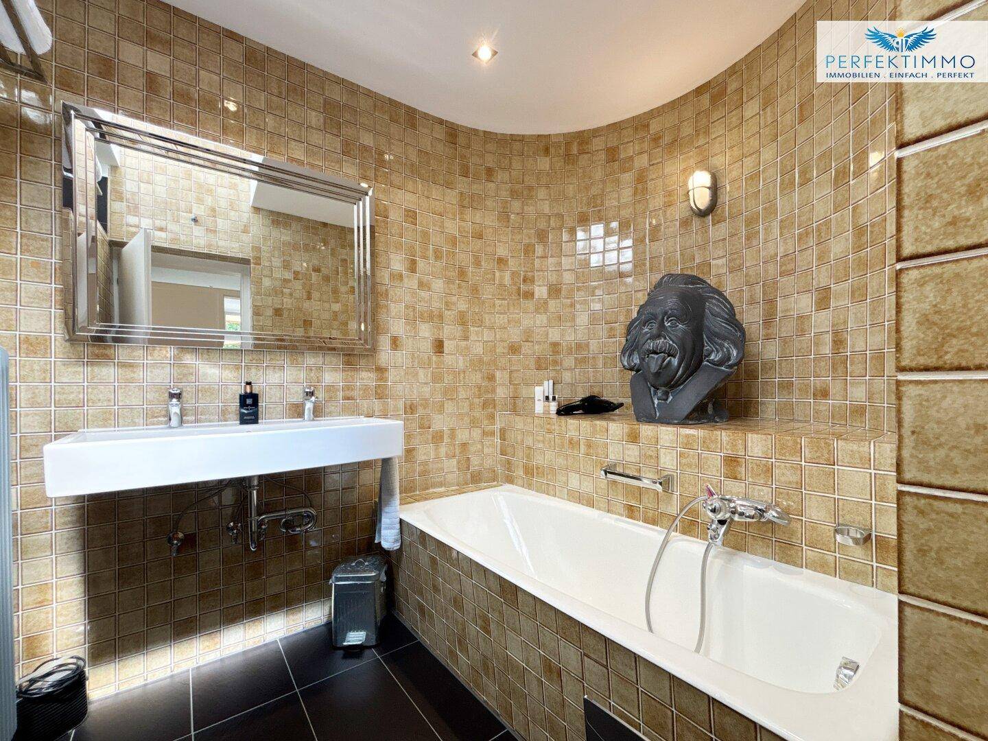 Badezimmer mit beigen Fliesen, einer Badewanne und einem breiten Waschbecken mit Spiegel.