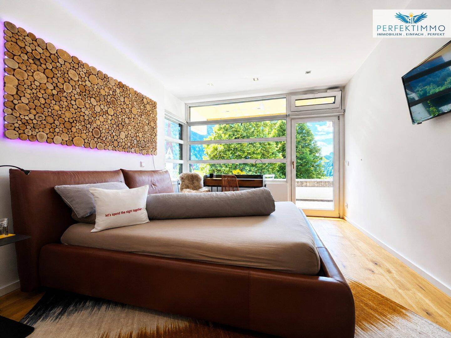 Helles Schlafzimmer mit braunem Bett, Holzboden und weitem Bergblick durch die Fensterfront.