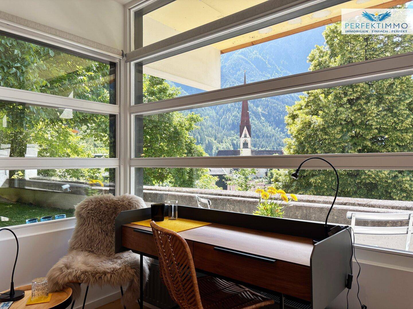 Arbeitsbereich mit großen Fenstern, die einen malerischen Blick auf Berge und eine Kirchturmspitze bieten.