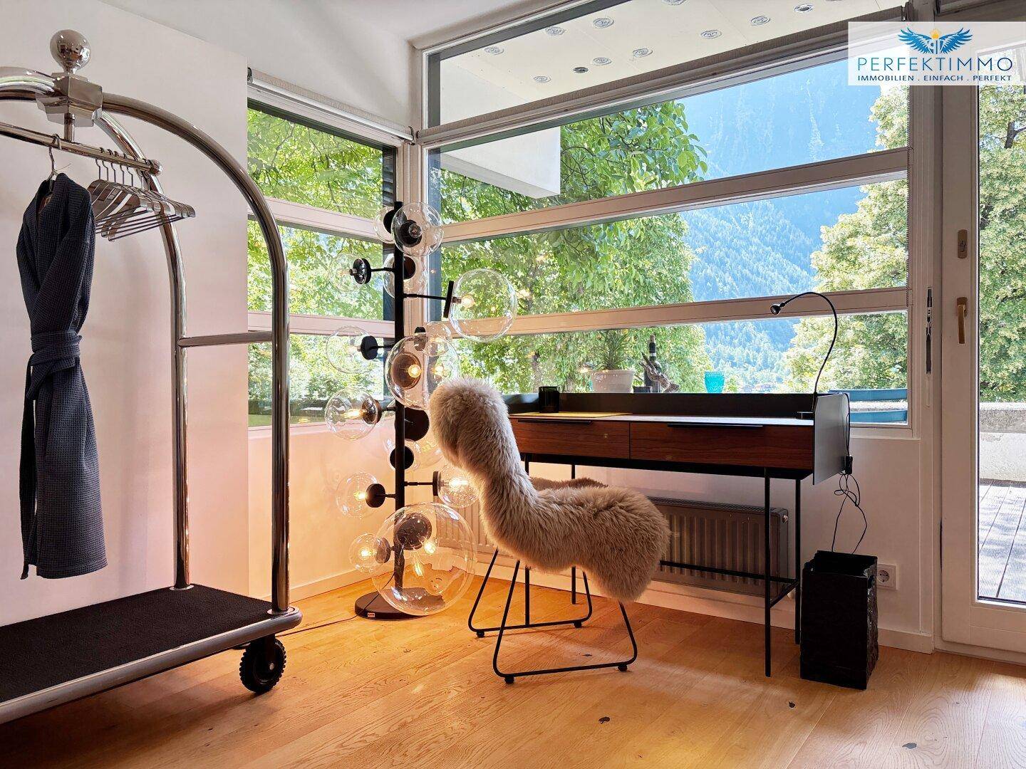 Heller Raum mit großen Fenstern, die einen grünen Ausblick bieten, und einem modernen Schreibtisch.
