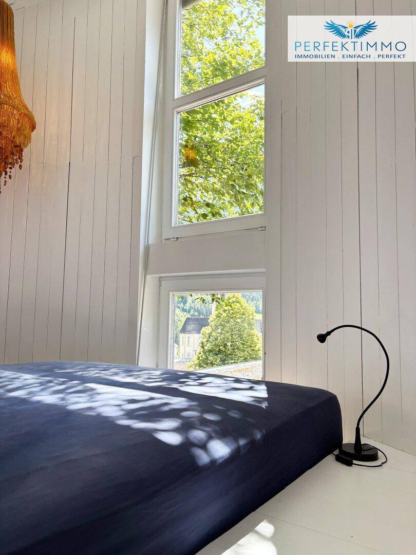 Helles Schlafzimmer mit hohen Fenstern und Blick auf die grüne Umgebung.