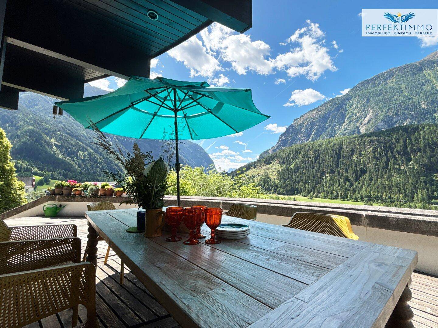 Sonniger Essbereich auf der Holzterrasse mit Bergblick und türkisfarbenem Sonnenschirm.