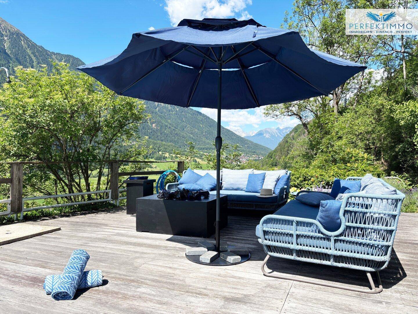 Gemütlicher Lounge-Bereich auf der Holzterrasse mit Sonnenschirm und Blick auf die Berge.