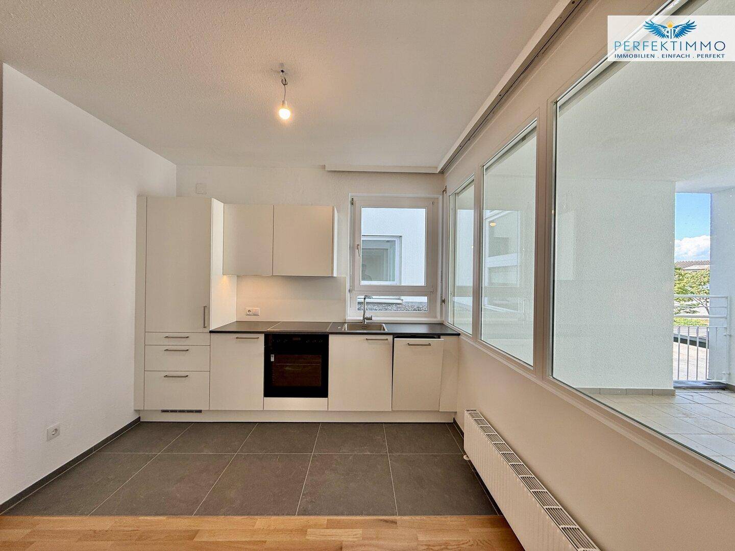 Moderne Einbauküche mit weißen Schränken, dunkler Arbeitsplatte und Fliesenboden, hell durch große Fenster.