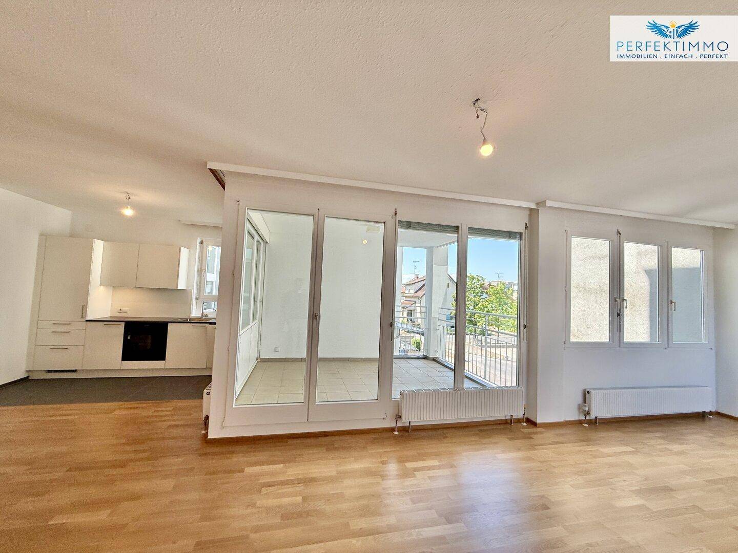 Geräumiges Wohnzimmer mit hellem Parkettboden und großen Fenstern, die viel Tageslicht hereinlassen.