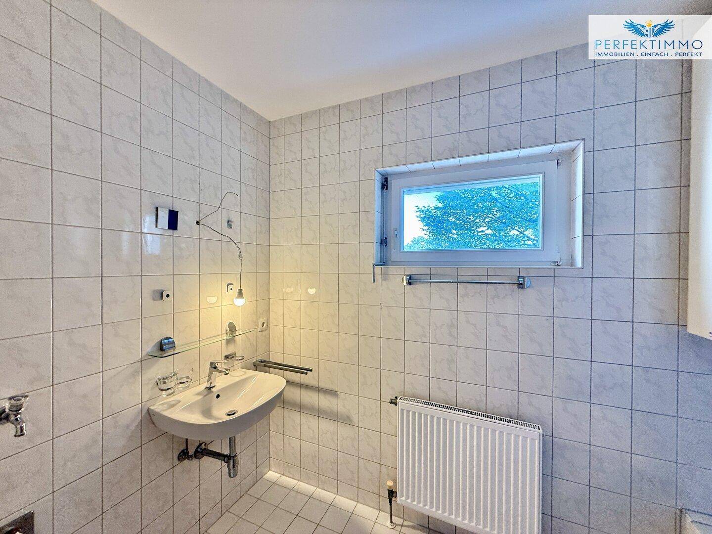 Badezimmer mit weißen Fliesen, Waschbecken und einem Fenster für natürliche Belüftung.