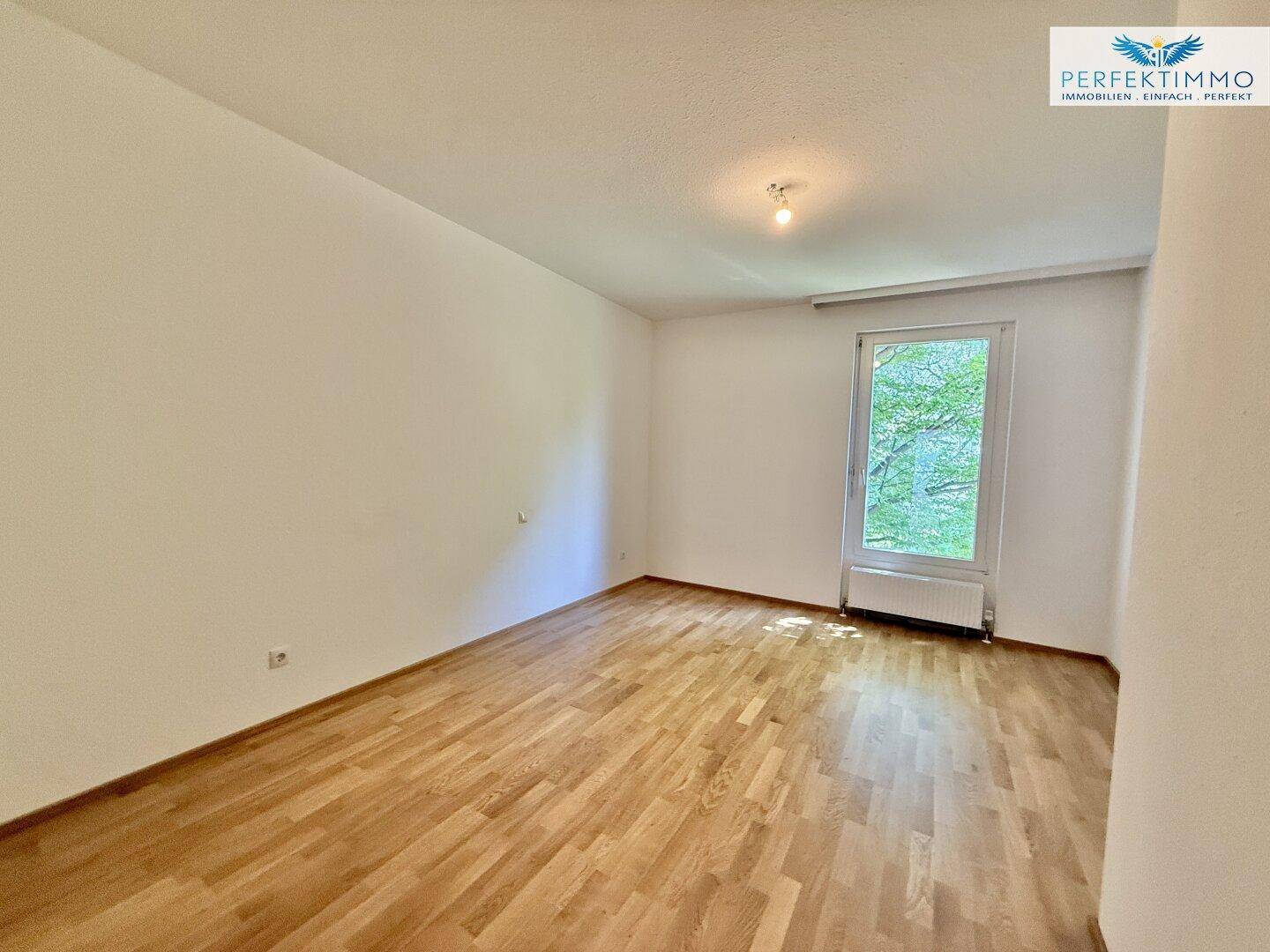 Helles Zimmer mit Parkettboden und einem Fenster, das einen Blick ins Grüne bietet.