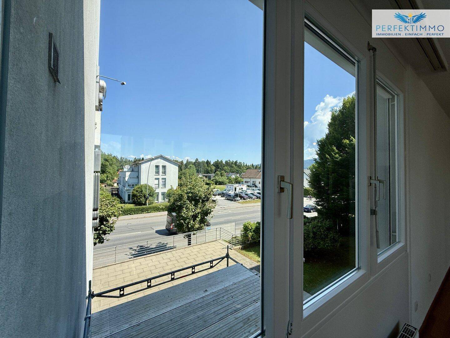 Ausblick aus dem Fenster auf die Straße, umliegende Gebäude und grüne Bäume.