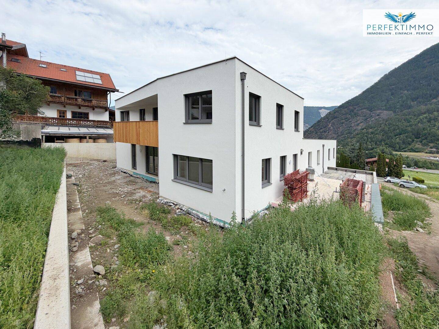 Neubau in alpiner Umgebung mit weißer Fassade und Holzakzenten, umgeben von viel Grün.
