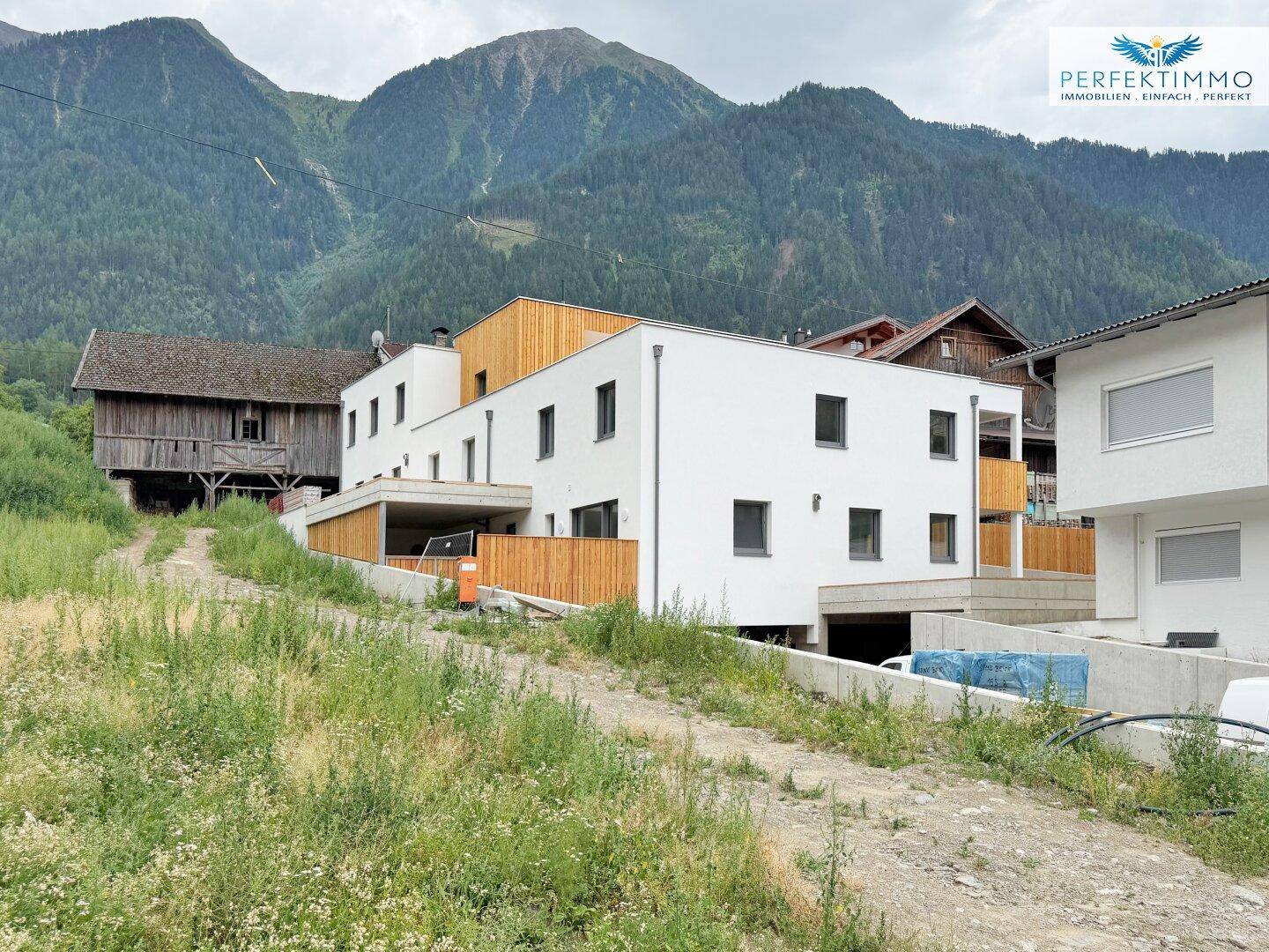 Modernes Gebäude mit weißer Fassade und Holzverkleidung vor alpiner Bergkulisse.