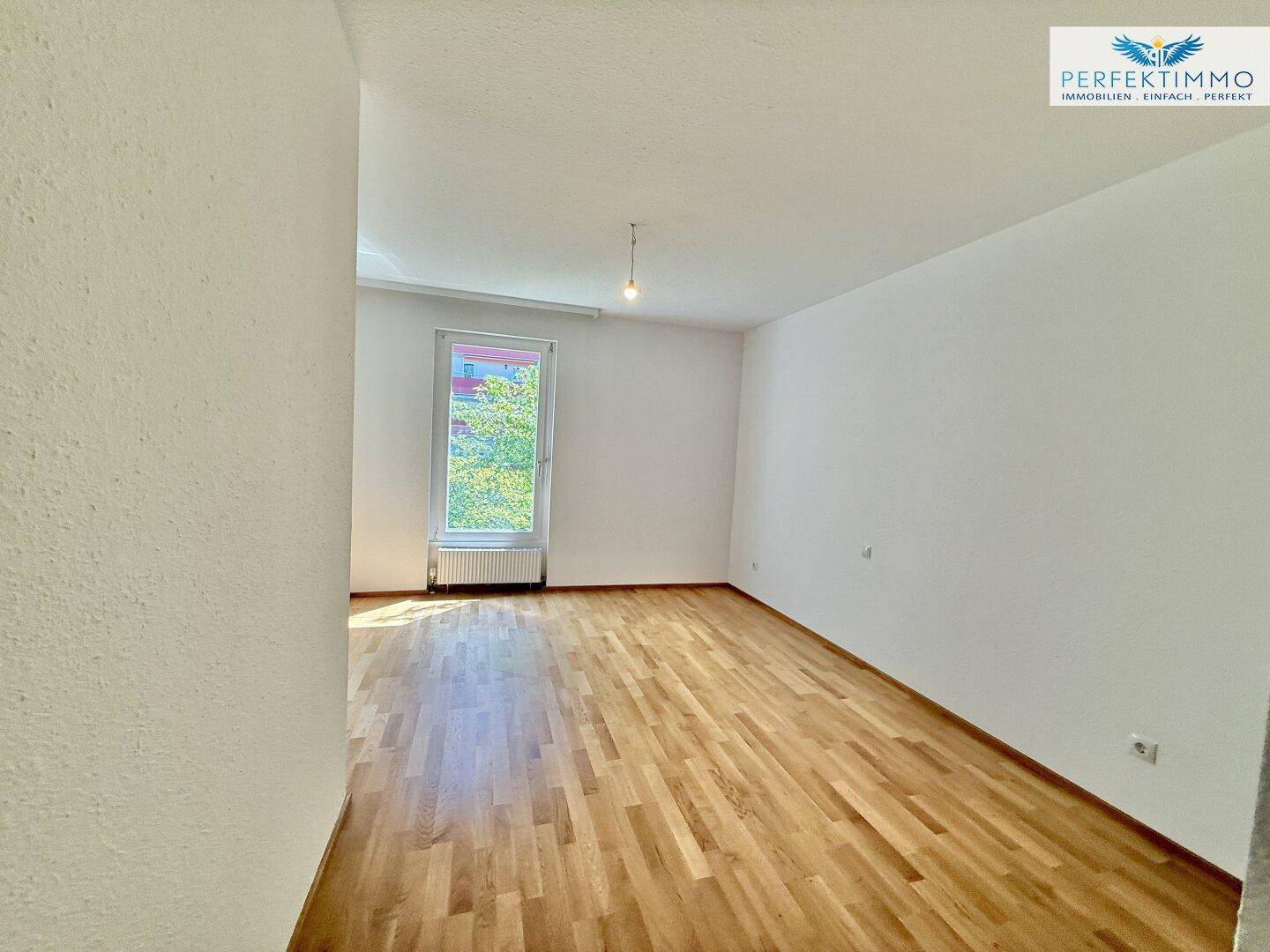 Geräumiges Schlafzimmer mit hellem Parkettboden und großem Fenster mit Grünblick.