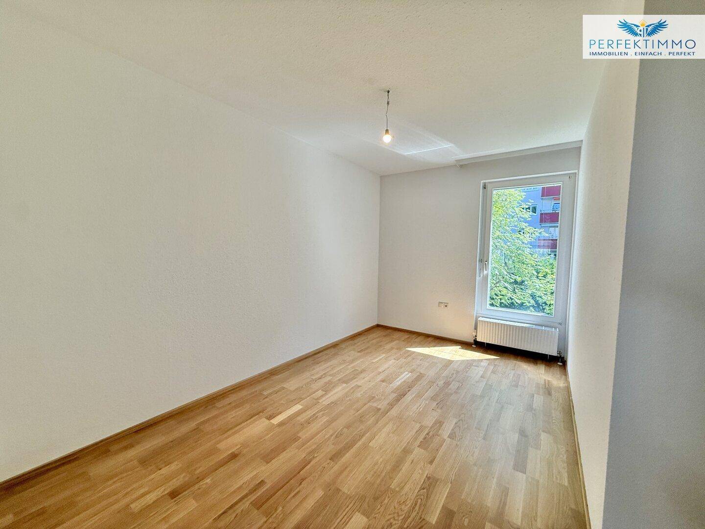 Helles Schlafzimmer mit Parkettboden und Fenster, das einen Blick ins Grüne bietet.