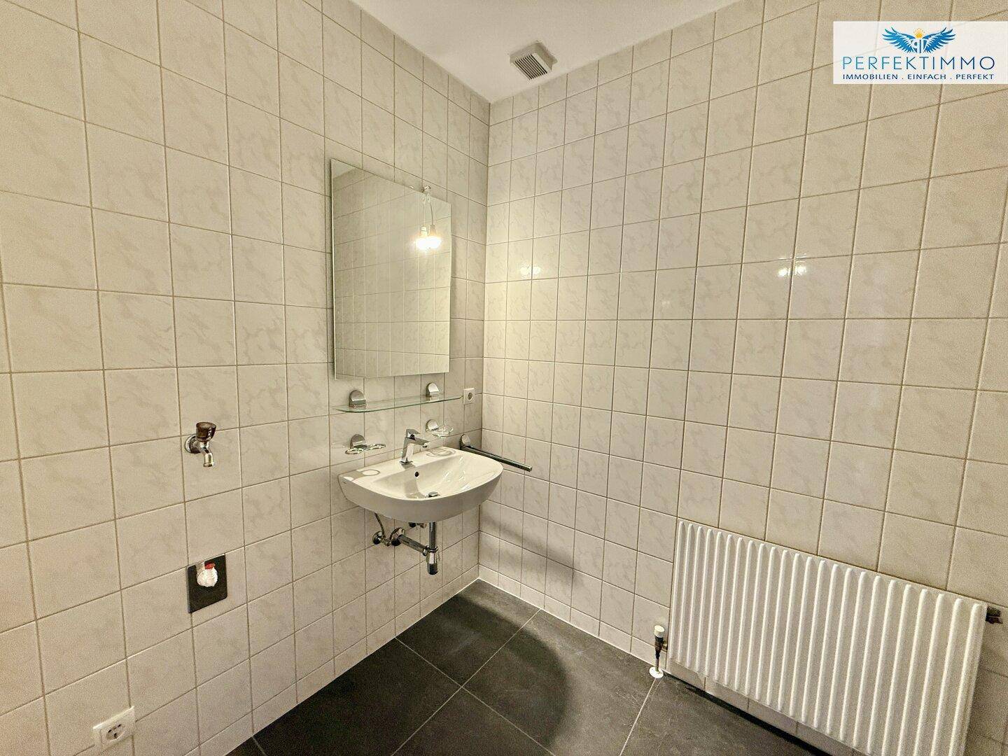 Gefliestes Badezimmer mit Waschbecken, Spiegel und Heizkörper, schlicht und funktional.