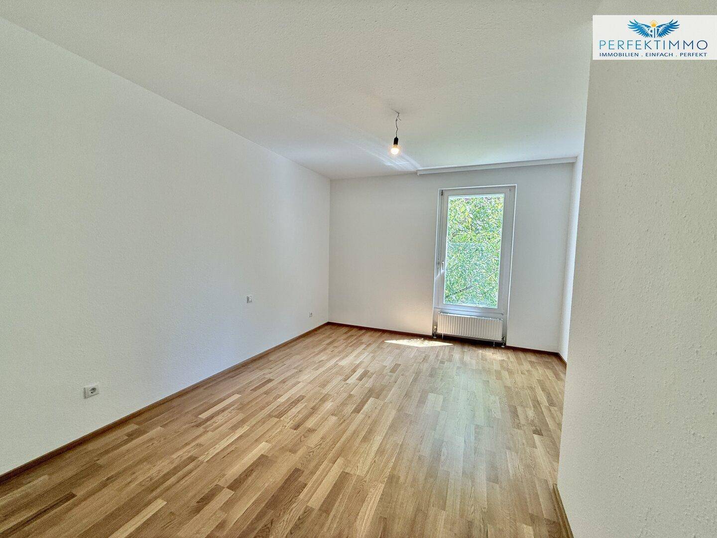 Helles Schlafzimmer mit Parkettboden und Fenster, das viel Tageslicht hereinlässt.