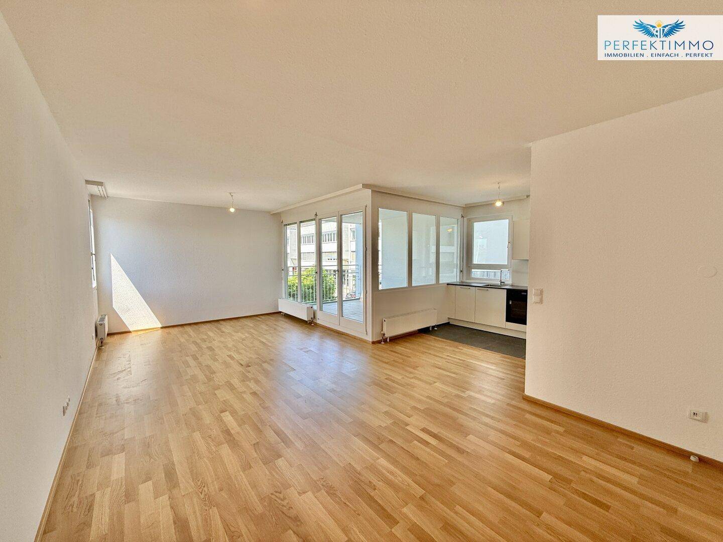 Geräumiger Wohnbereich mit hellem Holzboden, großen Fenstern und Zugang zum Balkon.
