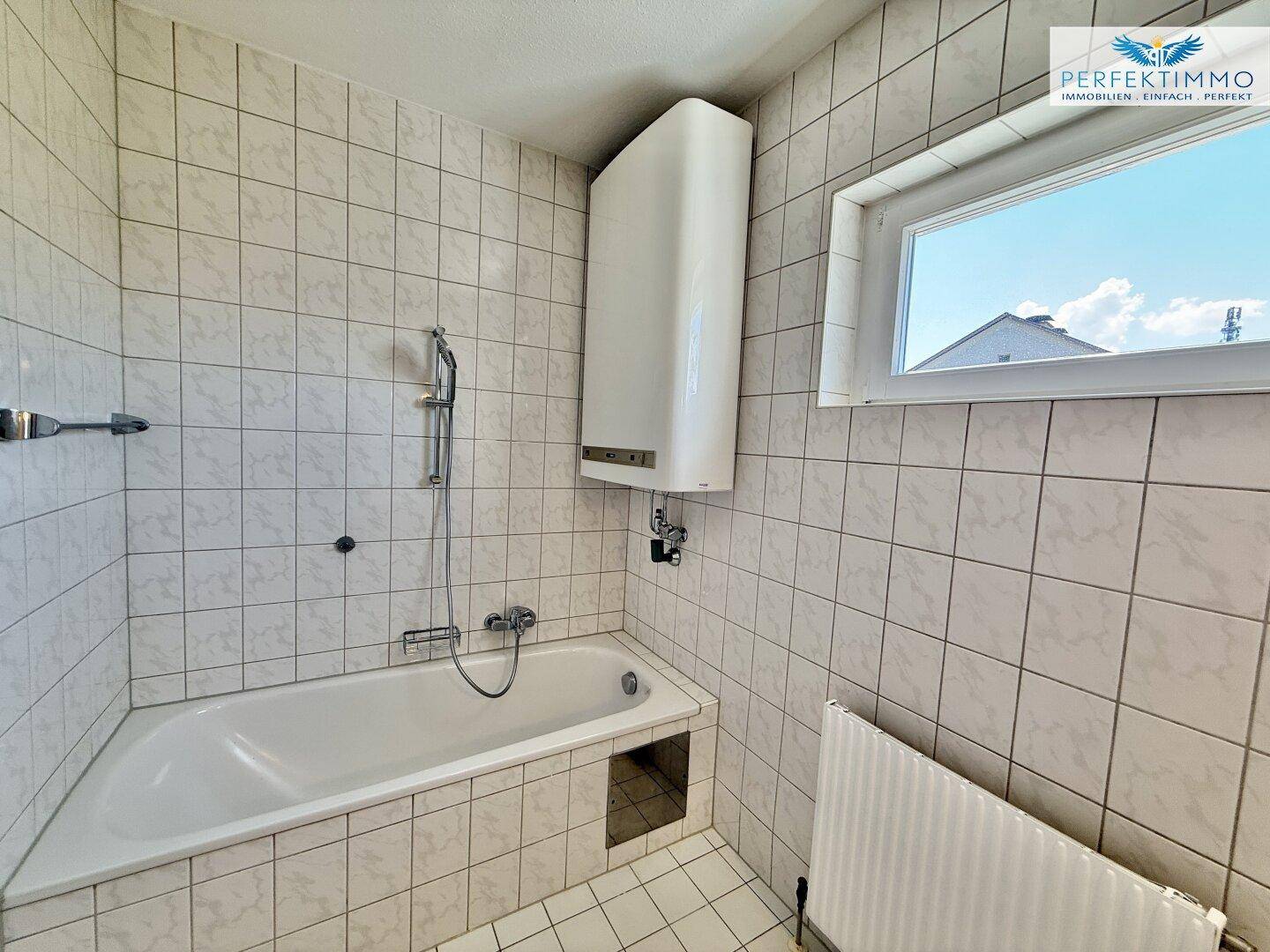 Gefliestes Badezimmer mit Badewanne, Wandheizung und Fenster für natürliche Belüftung.