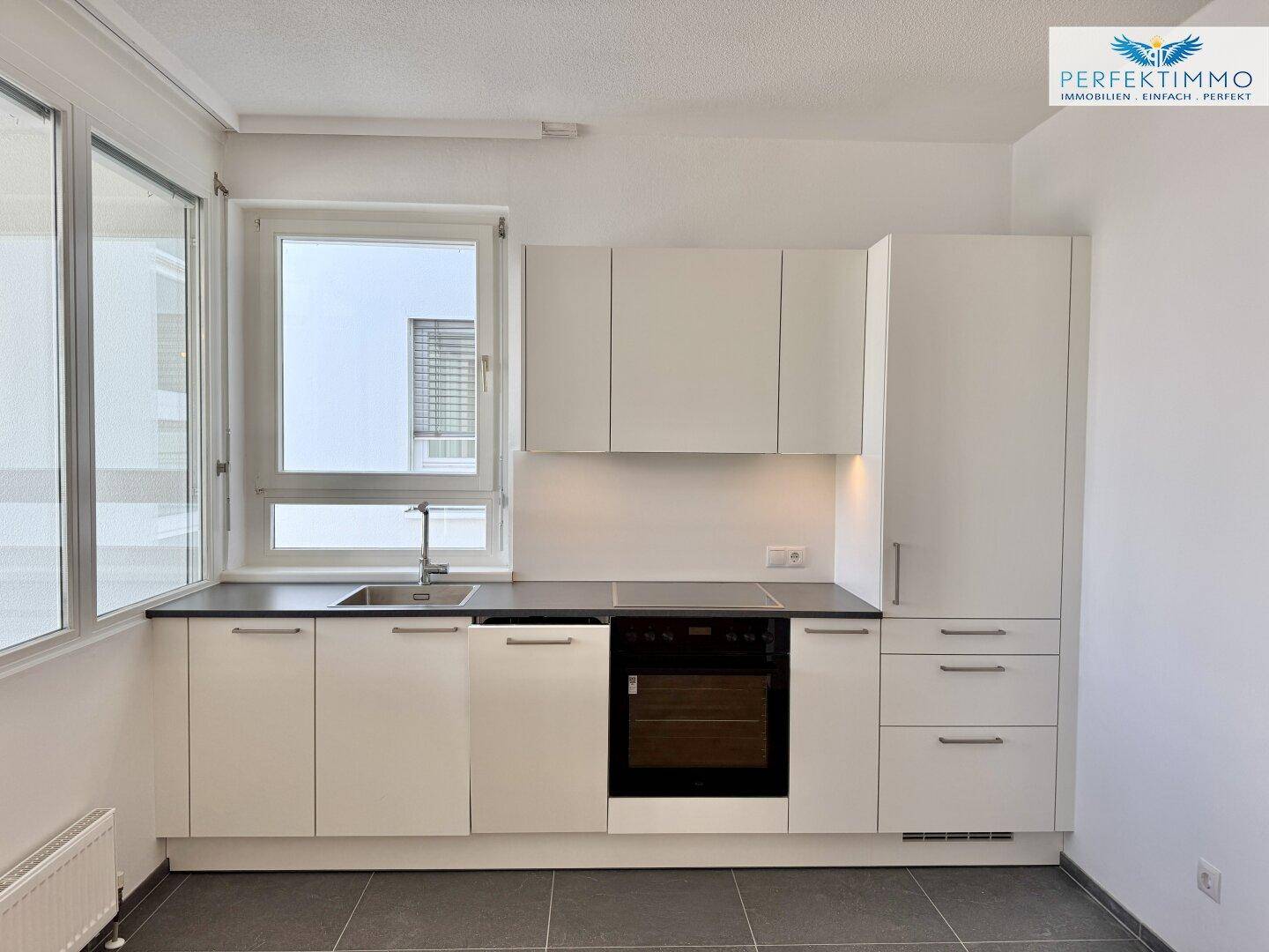 Moderne Einbauküche mit weißen Schränken, dunkler Arbeitsplatte und Fenster mit Ausblick.