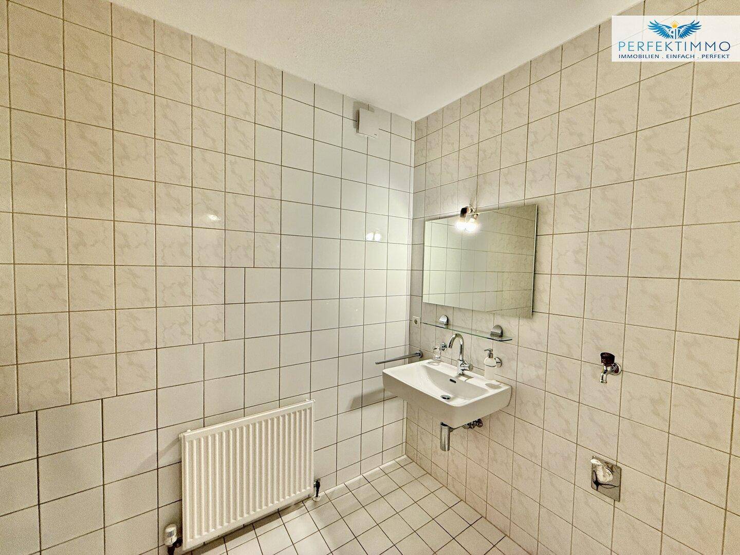 Gefliestes Badezimmer mit Waschbecken, Spiegel und Heizkörper, in hellem Zustand.