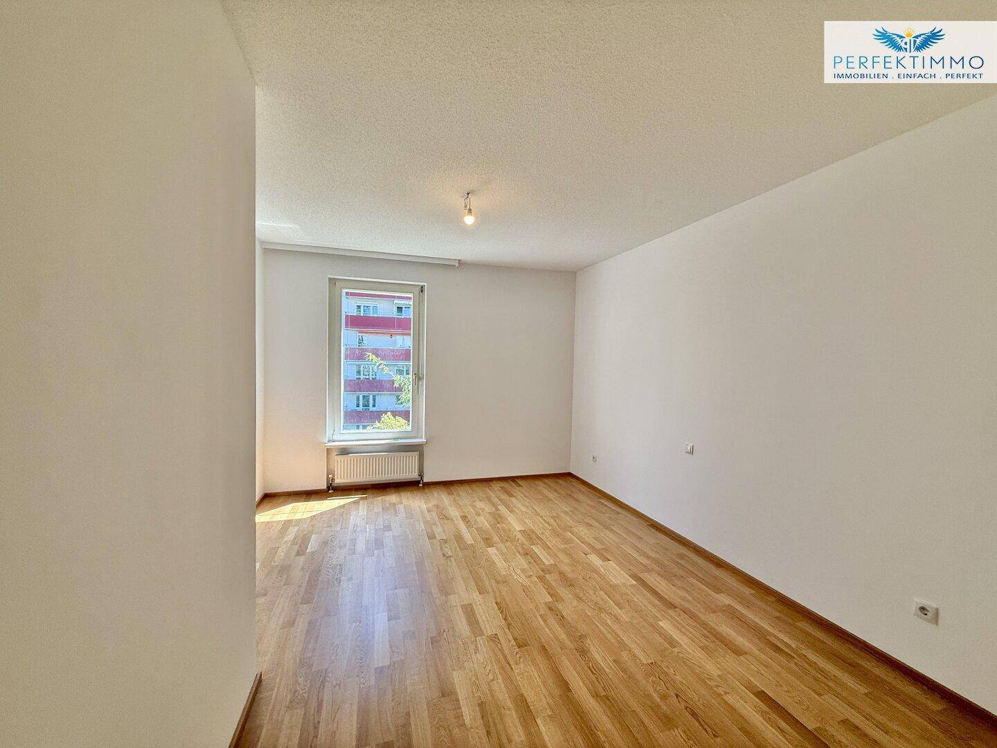Helles Schlafzimmer mit Parkettboden und Fenster, das viel natürliches Licht hereinlässt.