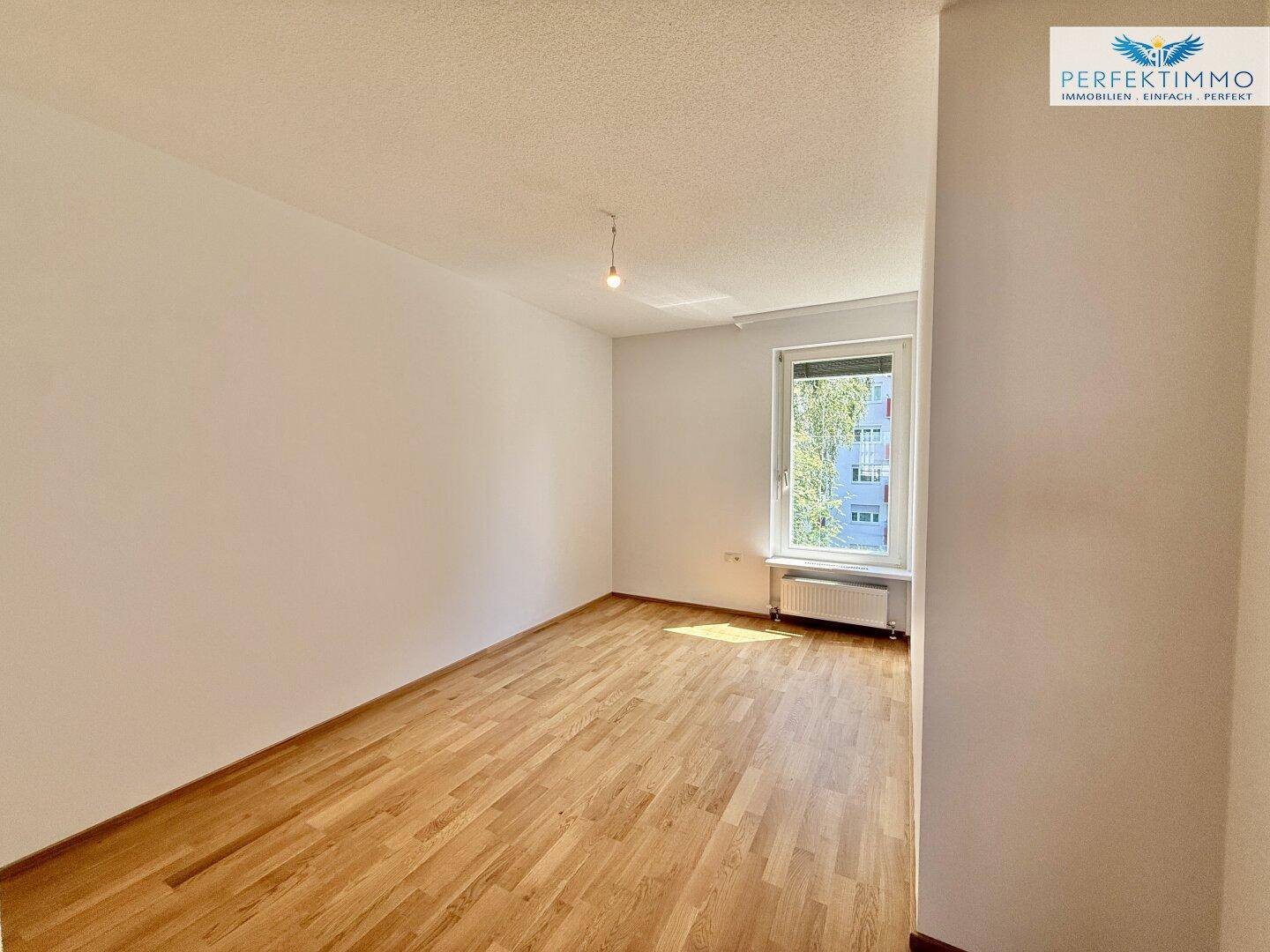 Helles Schlafzimmer mit Parkettboden und Fenster, das viel natürliches Licht hereinlässt.