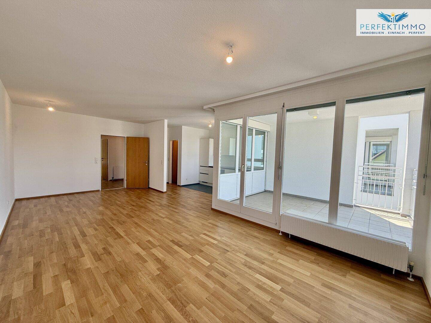 Weitläufiger Wohnbereich mit hellem Parkettboden und Zugang zu einem verglasten Balkon.