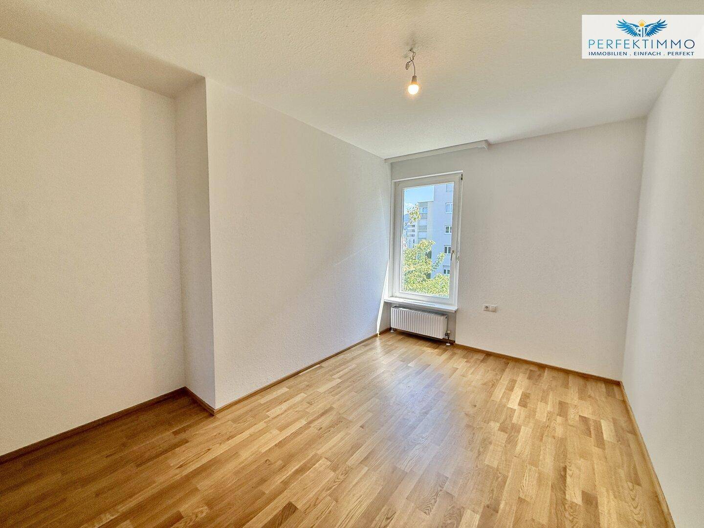 Helles Schlafzimmer mit Parkettboden und einem Fenster, das viel Tageslicht hereinlässt.