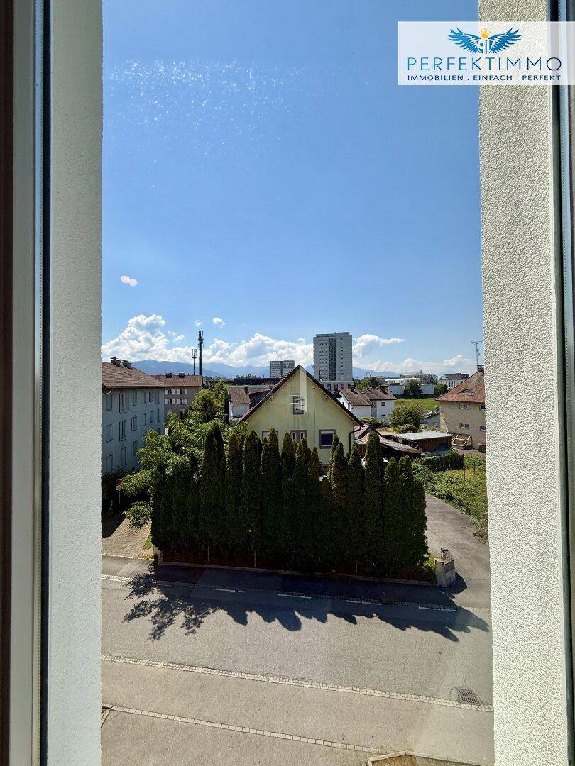 Weitblick aus dem Fenster über die Nachbarschaft mit Bäumen und fernen Bergen unter blauem Himmel.