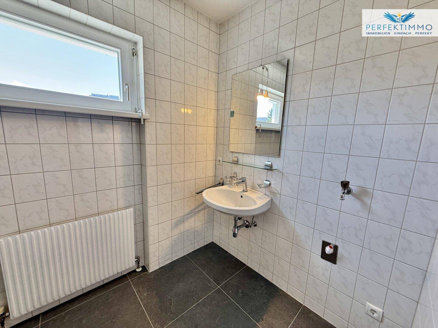 Gefliestes Badezimmer mit Waschbecken, Spiegel und Fenster, das für natürliche Belüftung sorgt.