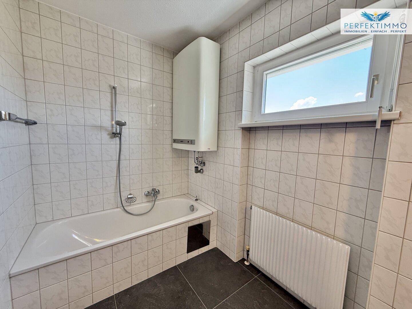 Badezimmer mit Badewanne, Fliesenboden und Fenster, ausgestattet mit einem Wandheizkörper.
