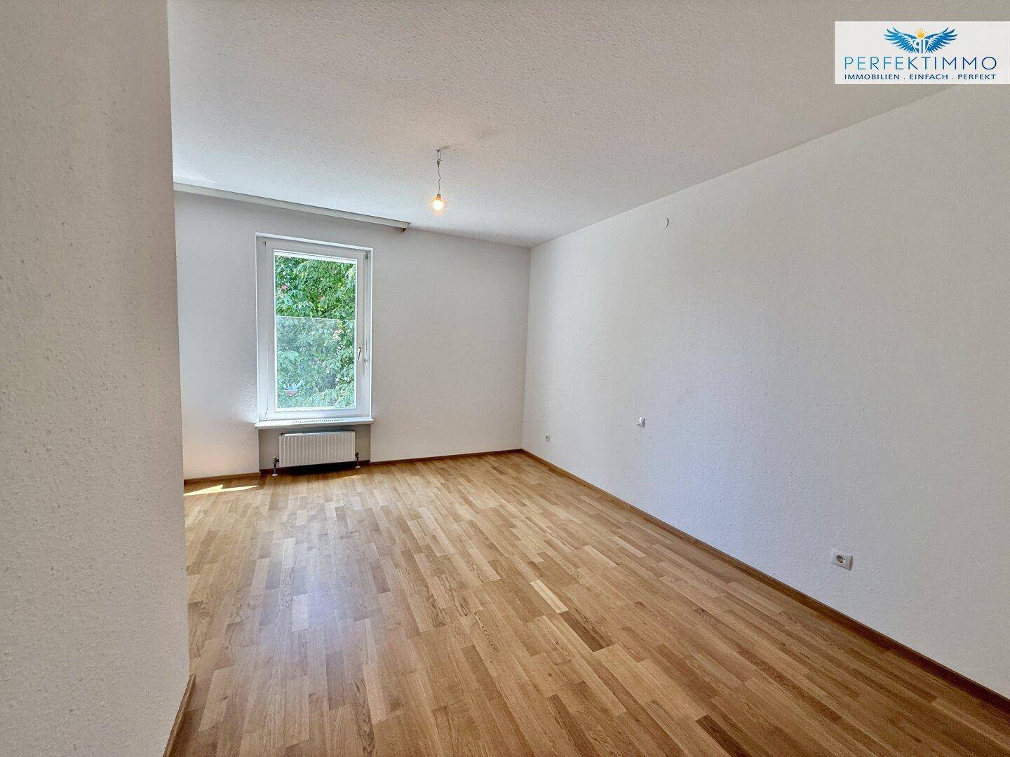 Helles Schlafzimmer mit Parkettboden, weißen Wänden und einem Fenster mit Blick ins Grüne.